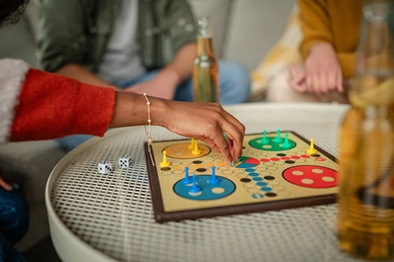 Hand moving a blue pawn on a classic Ludo board game placed on a coffee table with drinks nearby